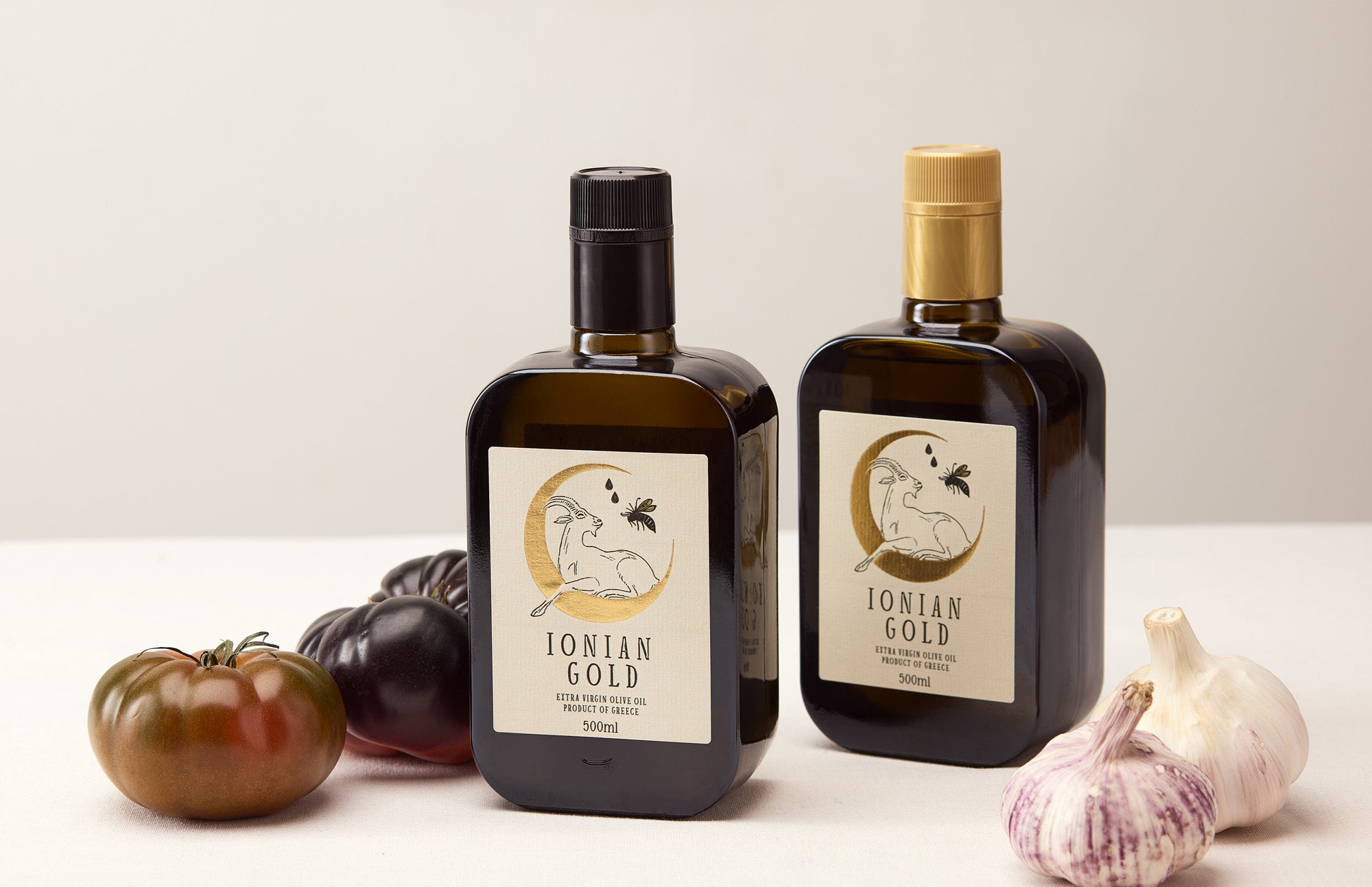 Two bottles of Ionian Gold'olive oil with vegetables on a white surface.