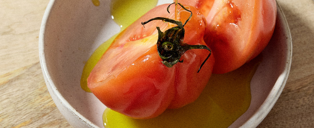 A close-up of a peeled and halved tomato resting in a white bowl with a small pool of golden olive oil on a wooden surface.