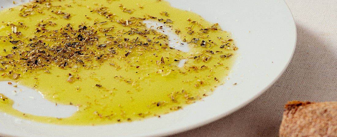 Close-up of a white plate with extra virgin olive oil drizzled and sprinkled with dried herbs, next to a piece of crusty bread on a light-colored surface.