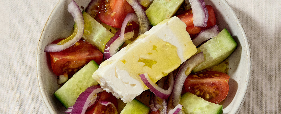 Bowl of Greek salad with cucumber slices, tomato wedges, red onion rings, topped with a rectangular slice of feta cheese drizzled with olive oil on a beige fabric surface.